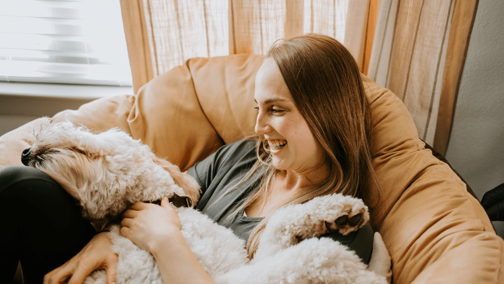 A smiling woman lounging on a cozy chair, holding and cuddling a fluffy dog in her lap.