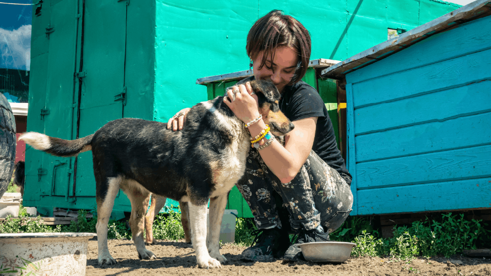 Person hugs a black-and-tan dog outside by turquoise buildings.