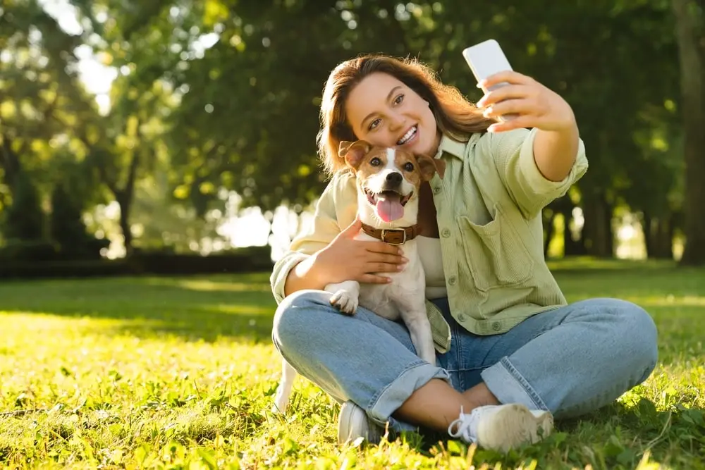 A Jack Russell terrier leans over a young woman's who sits cross-legged on grass outdoors to take a selife with a phone.