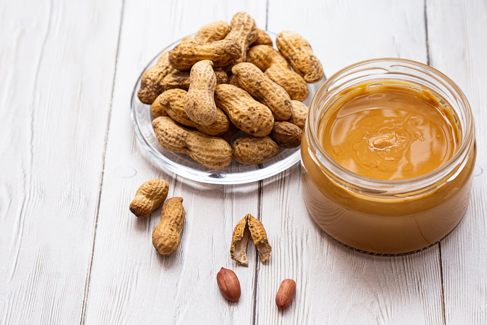 A glass jar of peanut butter with peanuts on the table.