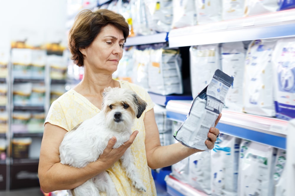 A woman reading a bag of dog food with a small dog in her arms. 