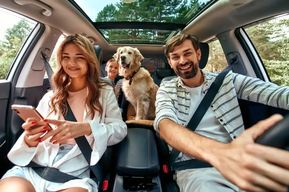 A happy family sits in a car with their child and dog in the backseat.