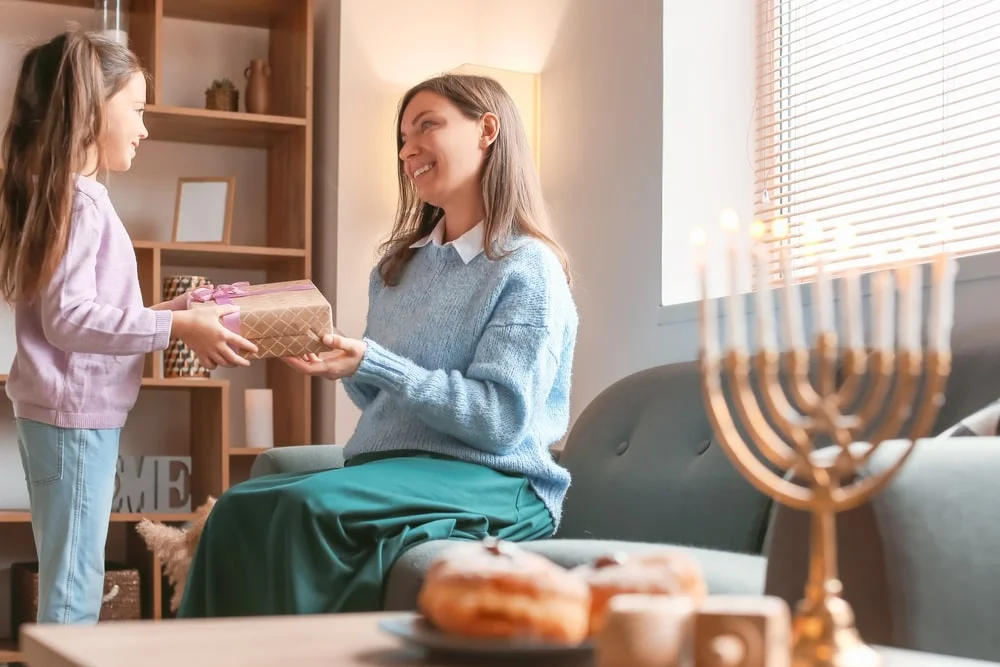 A parent and child exchange gifts with a menorah in the foreground.
