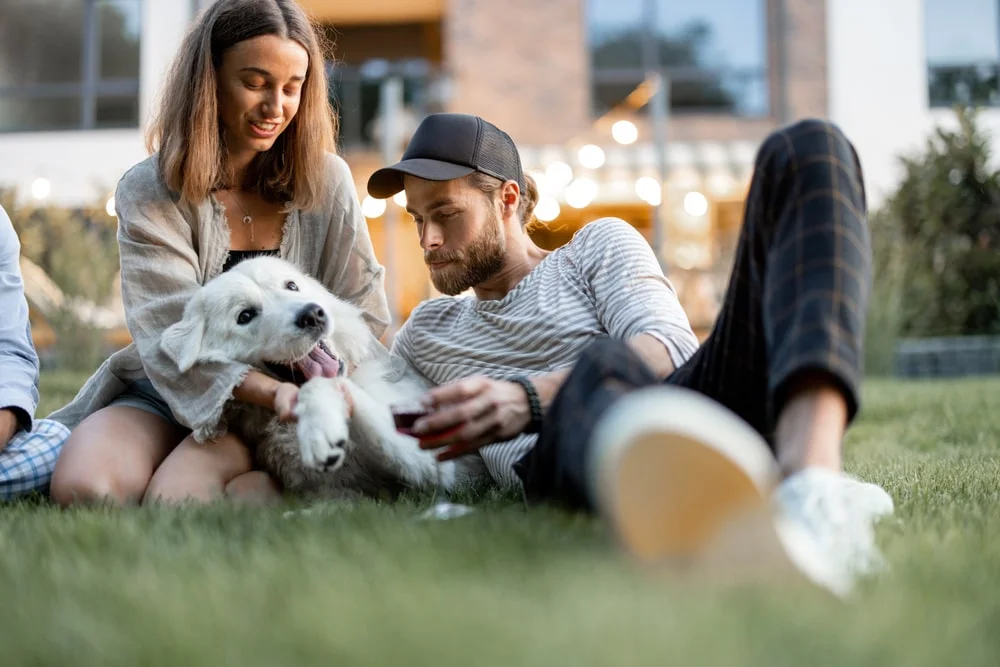 Two young people recline on grass with a white dog lounging between them.