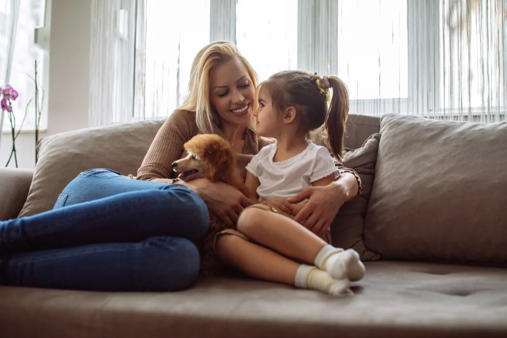 A parent and child sit on a couch with a brown poodle between them.