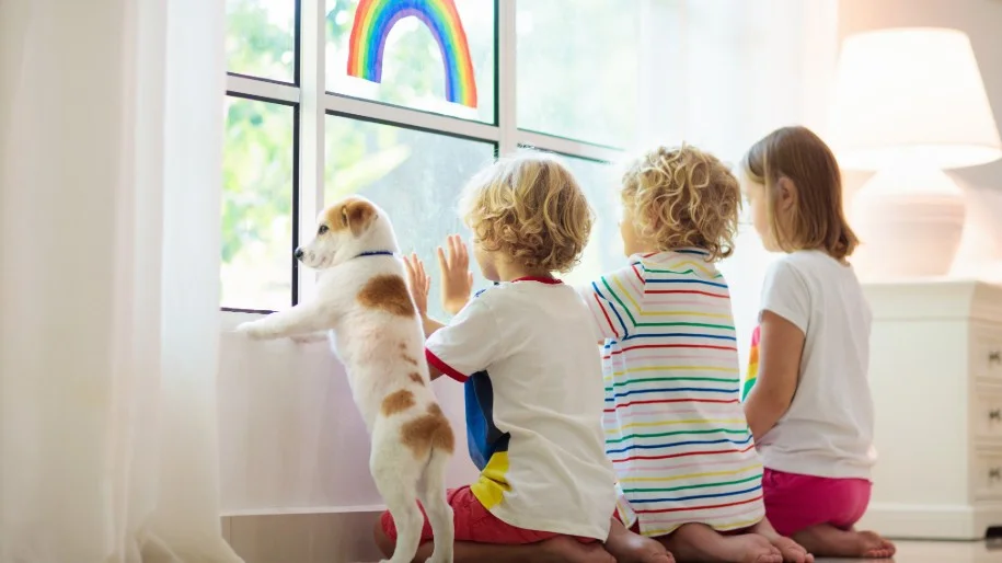 Three children and a brown and white puppy peer through a sunny window.